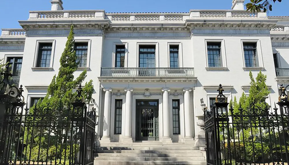 The image shows a grand white neoclassical building with a columned entrance iron gate and well-manicured landscaping