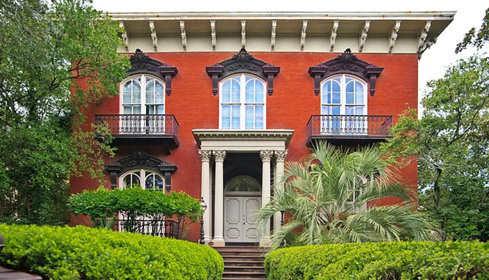 The image depicts a stately two-story red brick house with decorative black ironwork arched windows on the upper floor a grand entryway and lush greenery around it