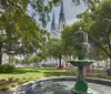 This image shows a serene urban park setting with lush trees a fountain in the foreground and a tall spire of a cathedral in the background under a clear blue sky