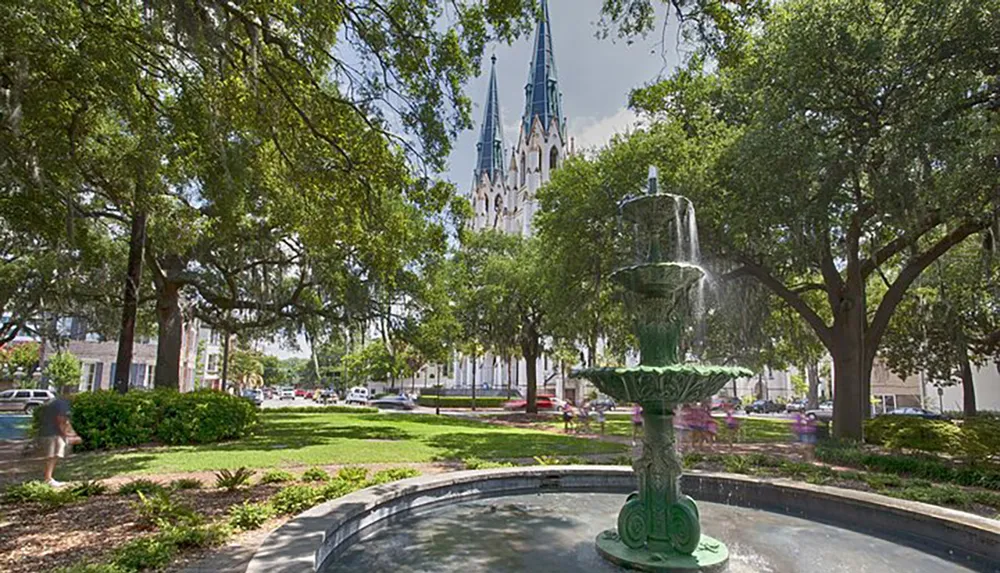 This image shows a serene urban park setting with lush trees a fountain in the foreground and a tall spire of a cathedral in the background under a clear blue sky