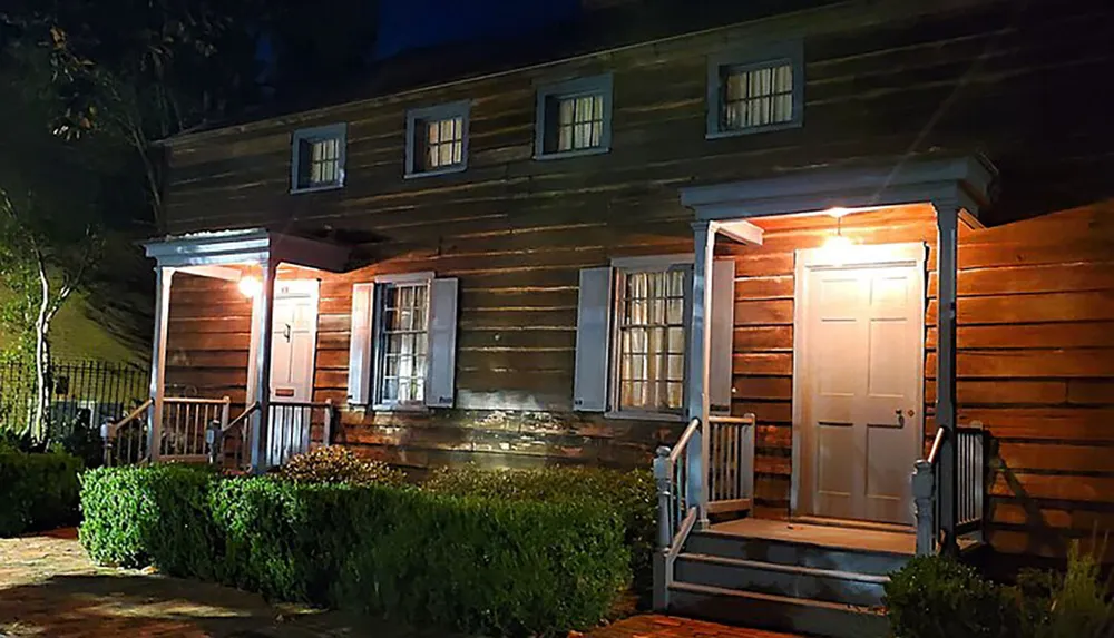 The image depicts a historic-looking wooden house illuminated by warm lighting at night featuring a porch with two entrances and white-trimmed windows