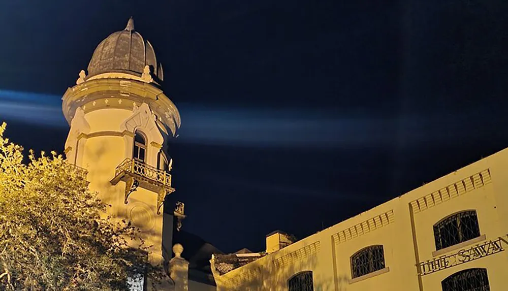 Illuminated by night lighting against a dark sky the image showcases an elegant dome-topped architectural turret connected to a building with classic details