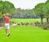 Two people are standing on a golf course with the person in the foreground holding a golf club looking towards the camera and the person in the background posing with a club over her shoulder