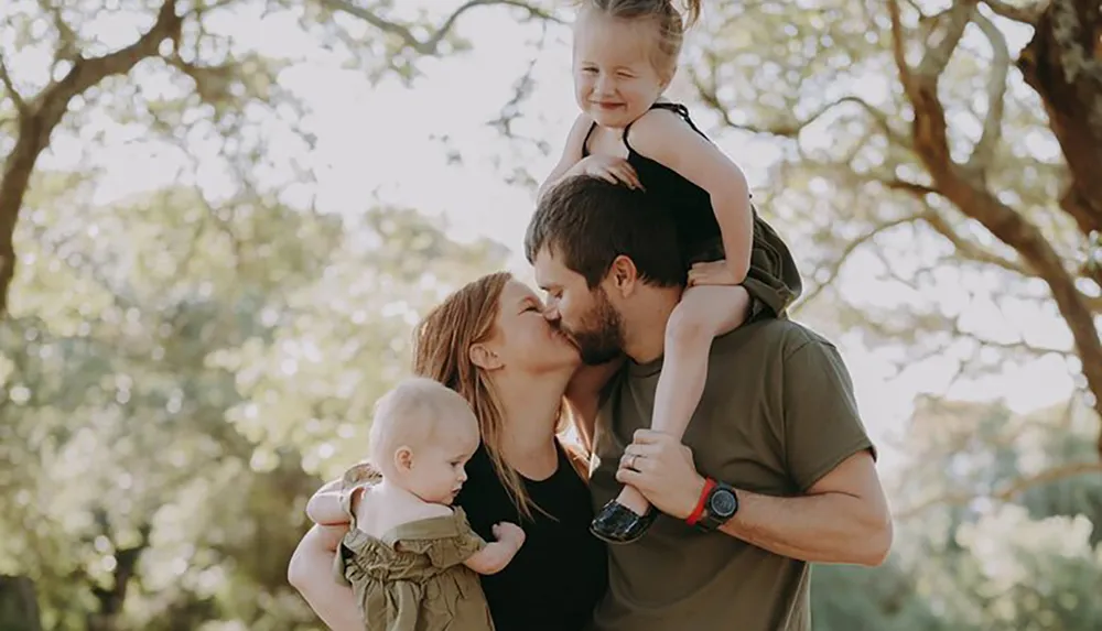 A happy family of four is enjoying a sunny outdoor moment with the parents kissing while each holds one of their young children