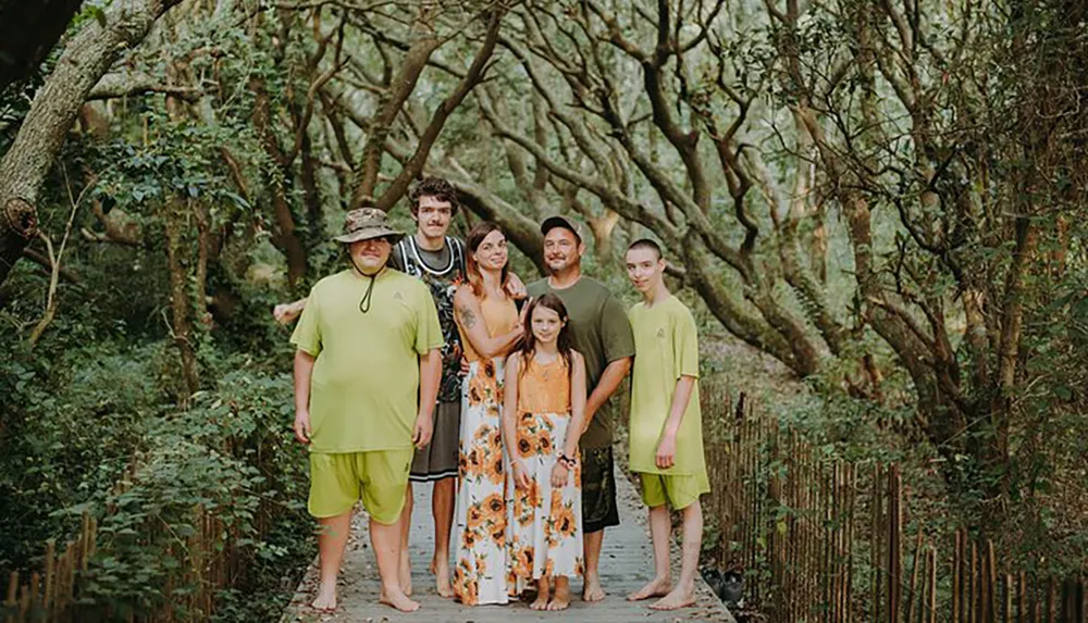 A family of six is posing for a photo on a wooded path flanked by lush greenery and winding tree branches