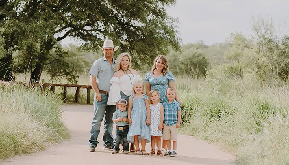 A family of seven is posing for a portrait on a sunny day on a country road with everyone dressed in coordinating blue outfits