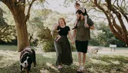 A family is enjoying a sunny day outdoors, with the parents holding their young children's hands and two dogs walking alongside.