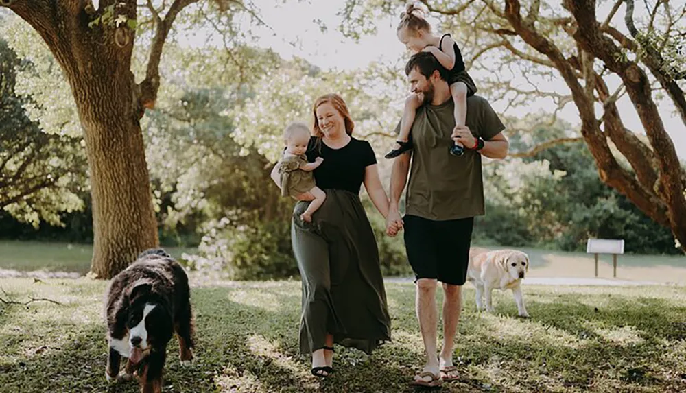 A family is enjoying a sunny day outdoors with the parents holding their young childrens hands and two dogs walking alongside