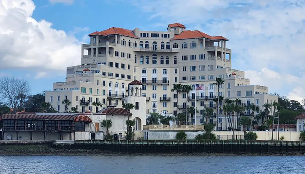 The image shows a multi-story palatial building with a white facade and terracotta roofs situated beside a body of water with palm trees and clear blue sky in the background