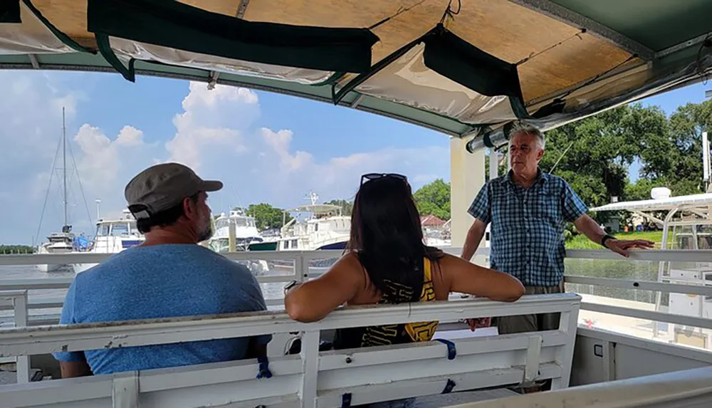 Three people are aboard a boat with a man standing and talking to a seated couple against a backdrop of a marina with various boats and a clear sky