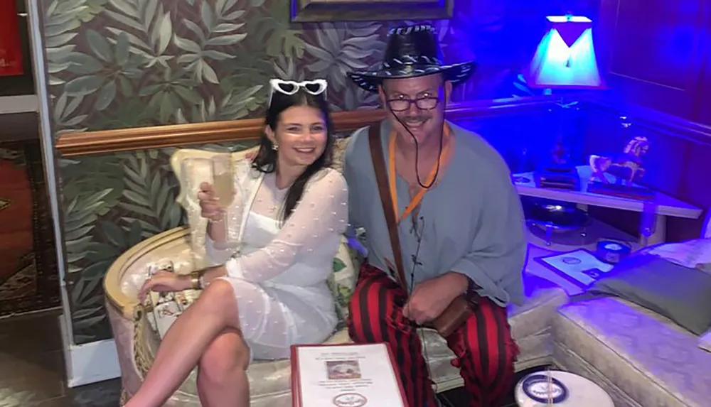 Two people are sitting and smiling for a photo in a room with stylish decor and colorful lighting with one person donning quirky sunglasses and the other wearing a hat and striped pants