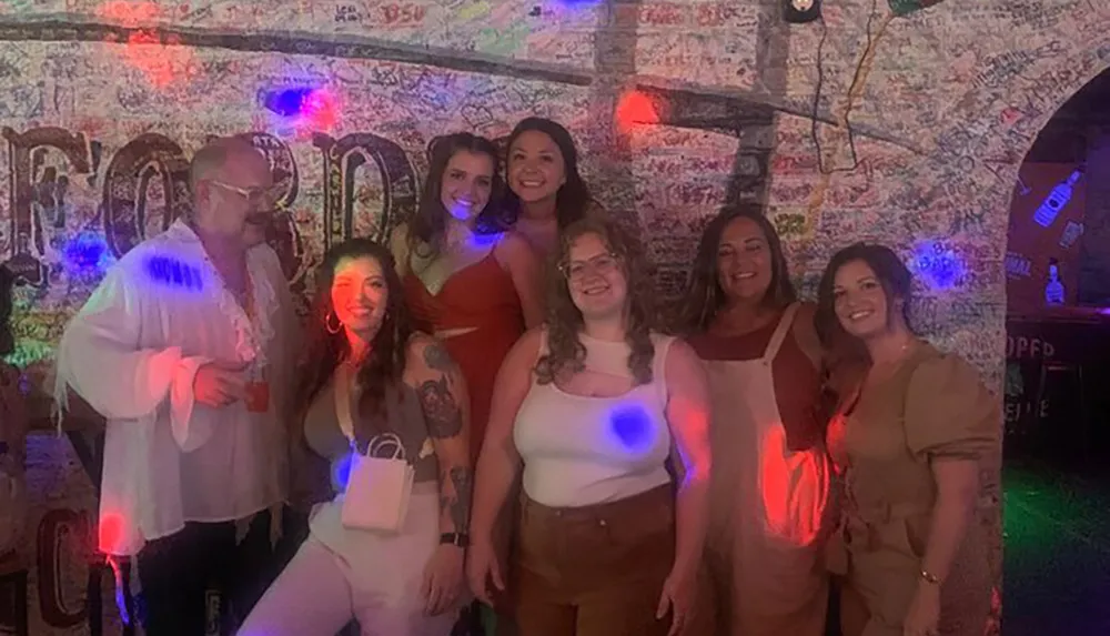 A group of seven people is posing for a photo in a room with vibrant lighting and a wall covered in graffiti