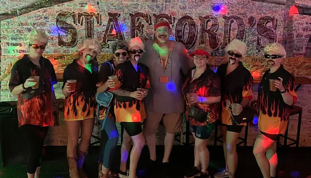 A group of people in matching flame-patterned outfits and white hair wigs several wearing sunglasses poses in front of a graffiti-covered wall holding drinks and smiling