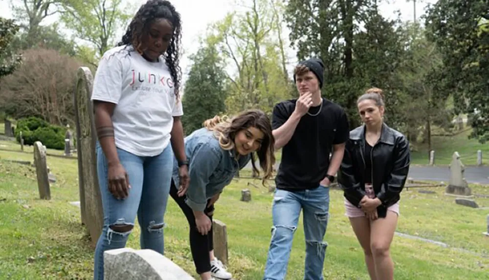 Four young adults stand pensively in a cemetery with some looking at a gravestone and others in contemplative poses