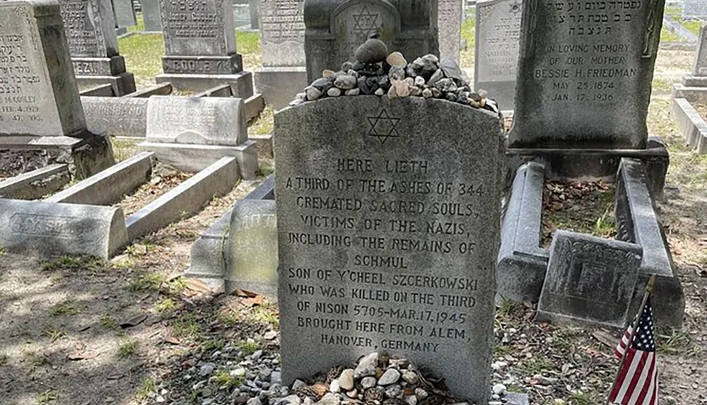 The image shows a gravestone marking the site where a third of the ashes of 344 cremated victims of the Nazis rest including specific mention of Schmul Szcerkowski with small stones placed on top in a Jewish mourning tradition and an American flag planted in front of it