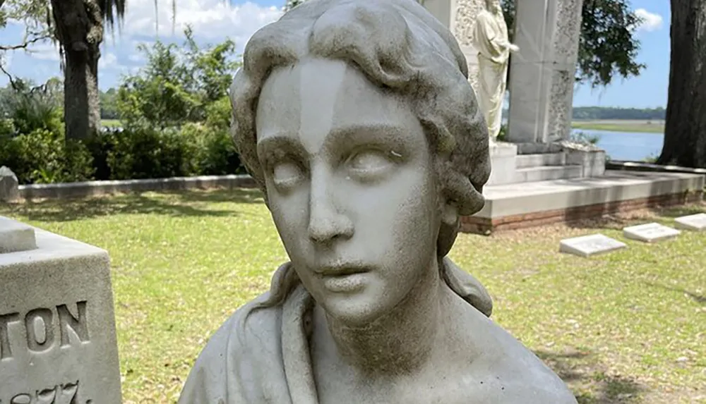 This is a weathered stone sculpture of a womans head and upper torso situated in a tranquil cemetery with headstones and trees in the background