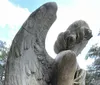 The image shows a sculpture of an angel with large detailed wings seated and looking contemplative against a backdrop of trees and a cloudy sky