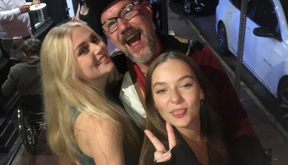 Three people are posing for a selfie on a street at night with two women smiling at the front and a man in a cap photobombing with a playful expression in the background