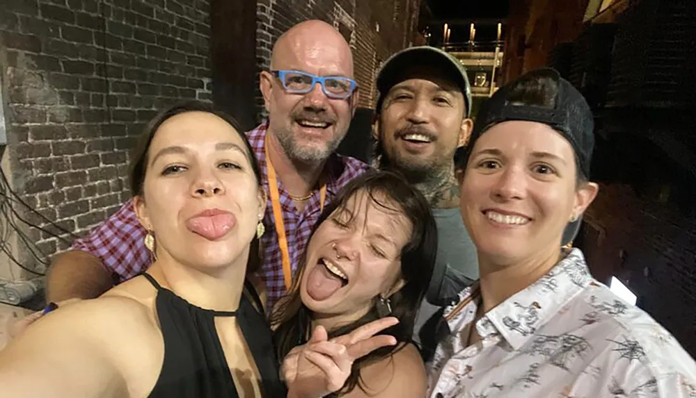 A group of five cheerful people are taking a nighttime selfie with some making playful faces and gestures