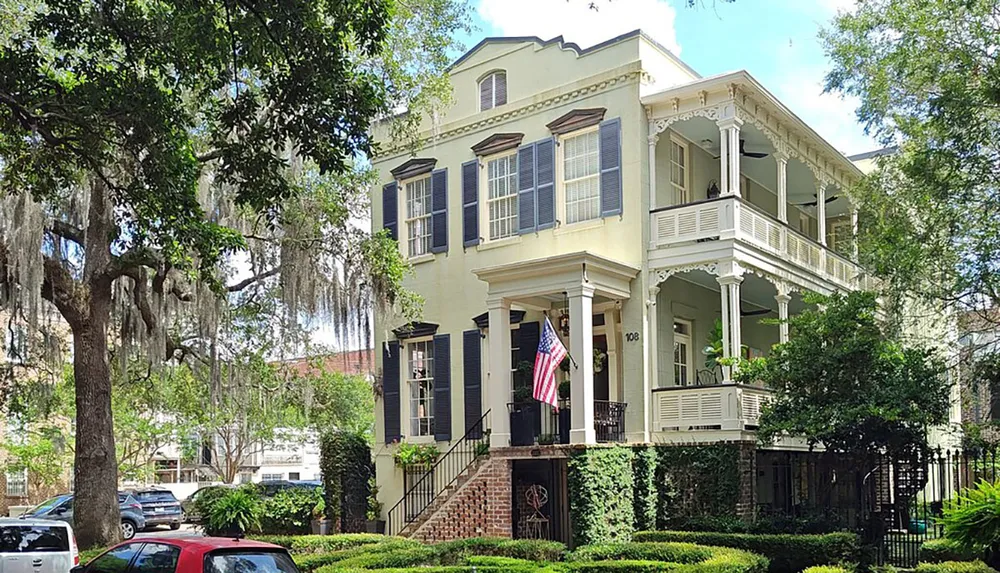 A charming two-story house with a double porch black shutters and an American flag is nestled among lush greenery under hanging Spanish moss