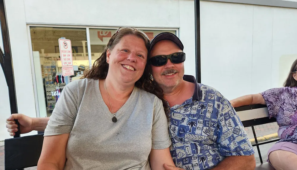 A smiling woman and a man wearing sunglasses are posing together seated on a bench with a storefront in the background