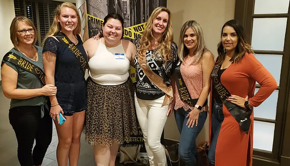 A group of six women smiling for the camera wearing sashes indicating a bride and her bridal party are at a celebration event