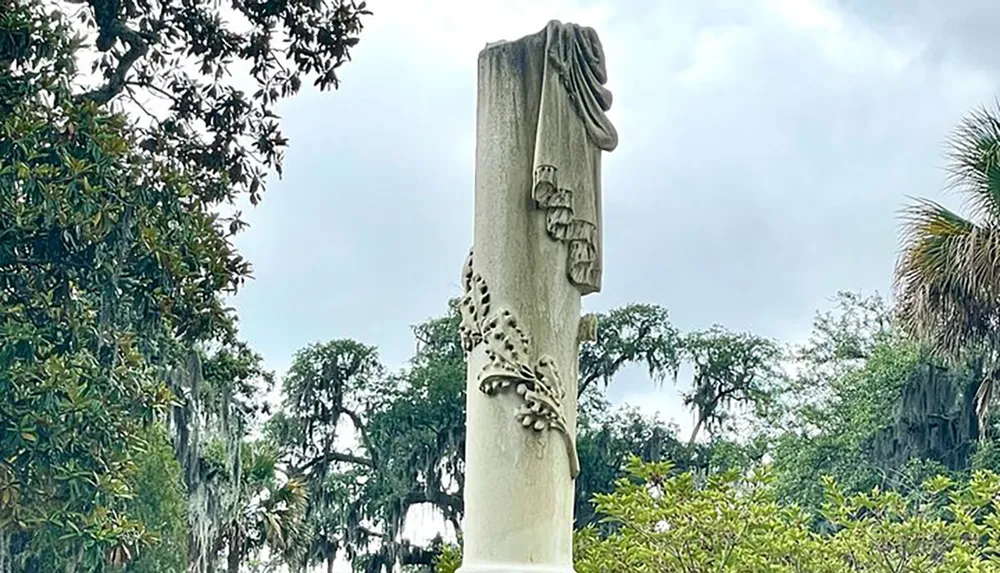 The image shows a unique columnar sculpture adorned with draped cloth and floral motifs set against a background of trees with hanging Spanish moss