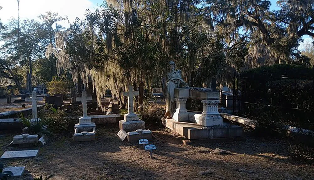 This image depicts an old cemetery with numerous tombstones and a statue amidst the trees draped with Spanish moss exuding a serene and somber atmosphere