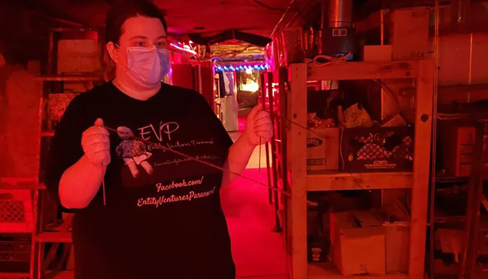 A person wearing a mask is standing in a dimly lit red-hued basement holding a dowsing rod