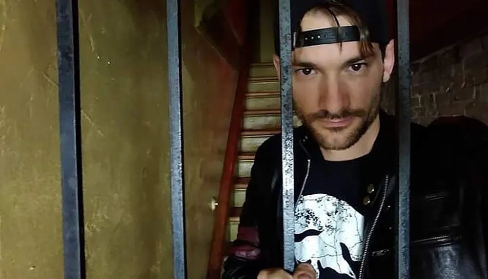 A man in a black cap and leather jacket is looking through iron bars with a staircase visible in the background