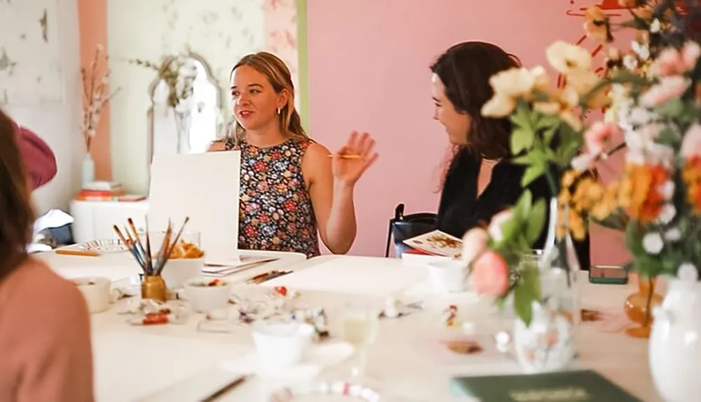 Two individuals are engaged in conversation at a table set up with art supplies and decorated with a colorful flower arrangement