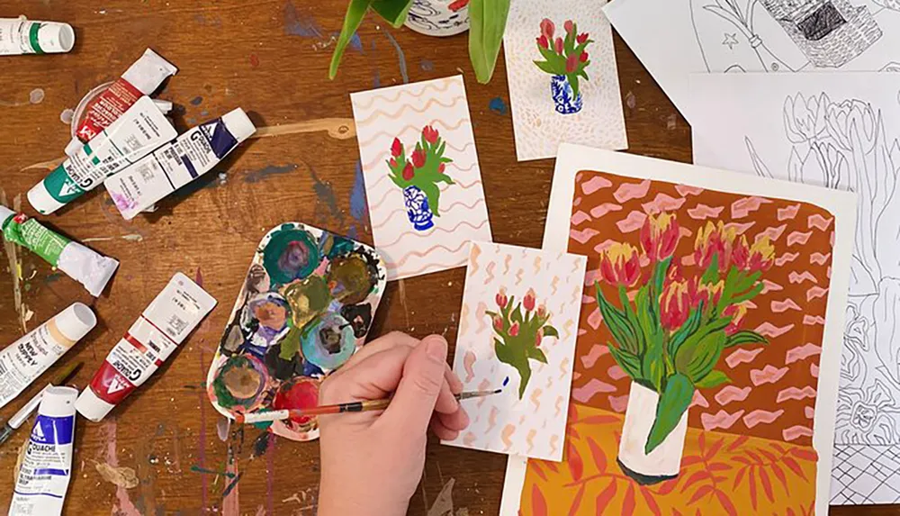 The image displays a painting activity in progress with various paintings of flowers an artists hand with a paintbrush and several tubes of paint on a messy worktable