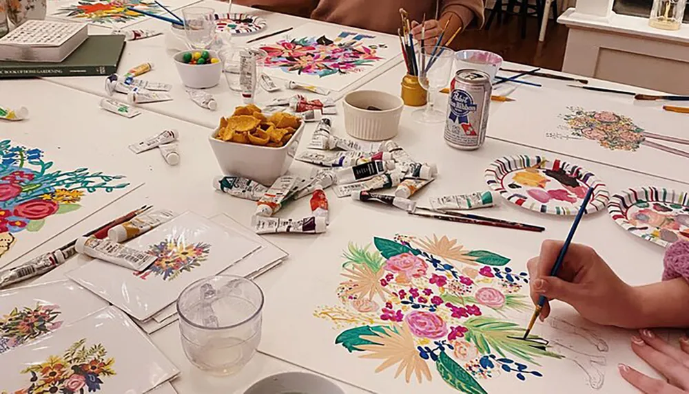 A persons hand is seen painting colorful floral patterns on a white table cloth amidst a creative setup with scattered paint tubes paintbrushes snacks and beverages