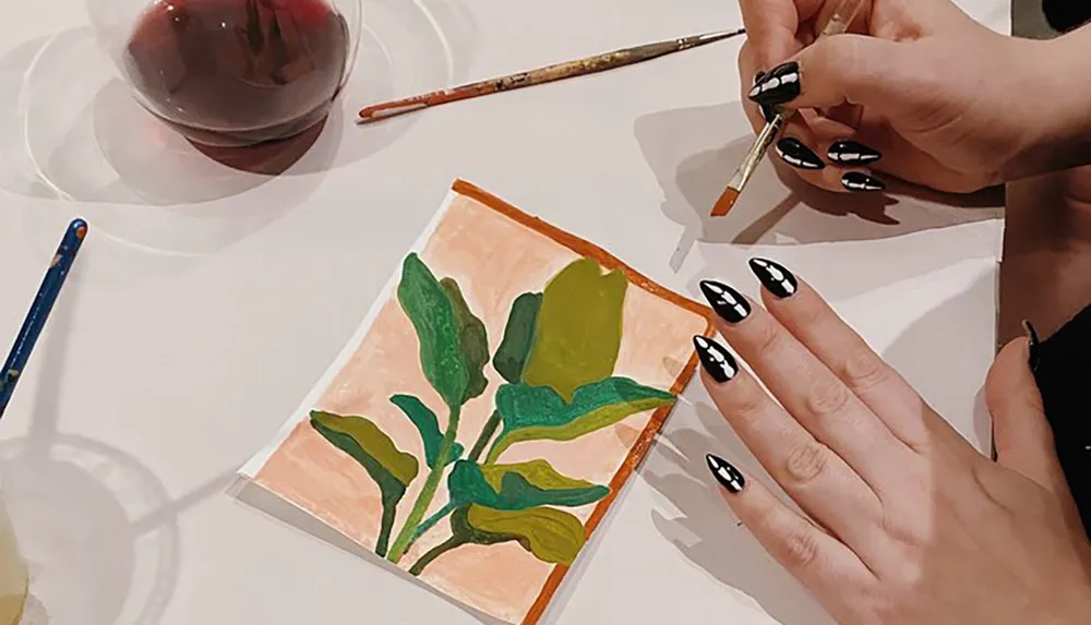 A person with creative nail art is painting a green plant on a small canvas on a white table next to a glass of red wine