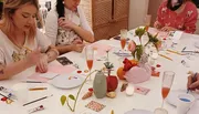 A group of people are engaged in a creative workshop, painting and crafting at a table decorated with colorful art supplies, flowers, and drinks.