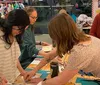 A group of people appears to be engaged in a craft or sewing workshop surrounded by fabric and sewing materials inside a well-lit room