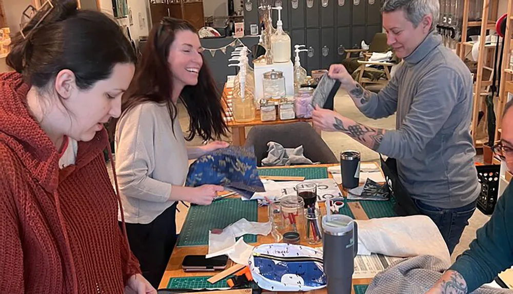 A group of people are engaged in what appears to be a craft workshop smiling and working on various projects around a table with supplies scattered across it