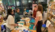 A group of people appears to be engaged in a craft or sewing workshop, surrounded by fabric and sewing materials, inside a well-lit room.