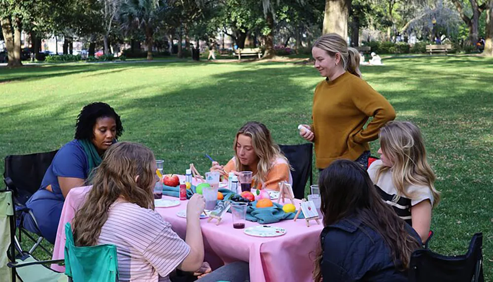 A group of people are gathered around a table in a park engaging in what appears to be an outdoor craft activity