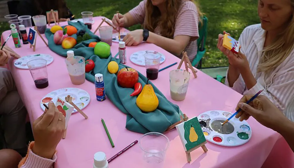 A group of individuals is engaged in an outdoor painting activity using a variety of fruits as subjects for their artwork