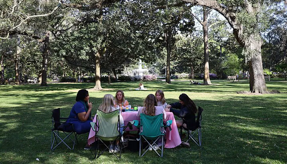 A group of people are enjoying a picnic at a table set up on a lush green lawn with trees and a fountain in the background