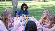 A group of people is enjoying a cheerful outdoor craft activity around a table covered in art supplies.