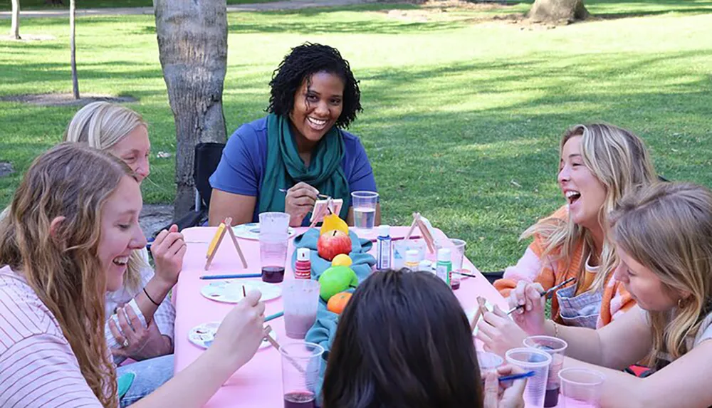 A group of people is enjoying a cheerful outdoor craft activity around a table covered in art supplies