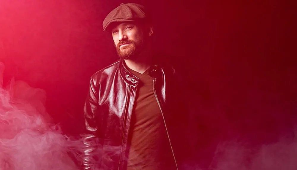 The image shows a bearded man wearing a flat cap and leather jacket set against a red-lit background with a hazy smoke effect