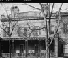 The image is a black and white photograph of a two-story historical building with a balcony surrounded by bare trees indicative of an older urban environment