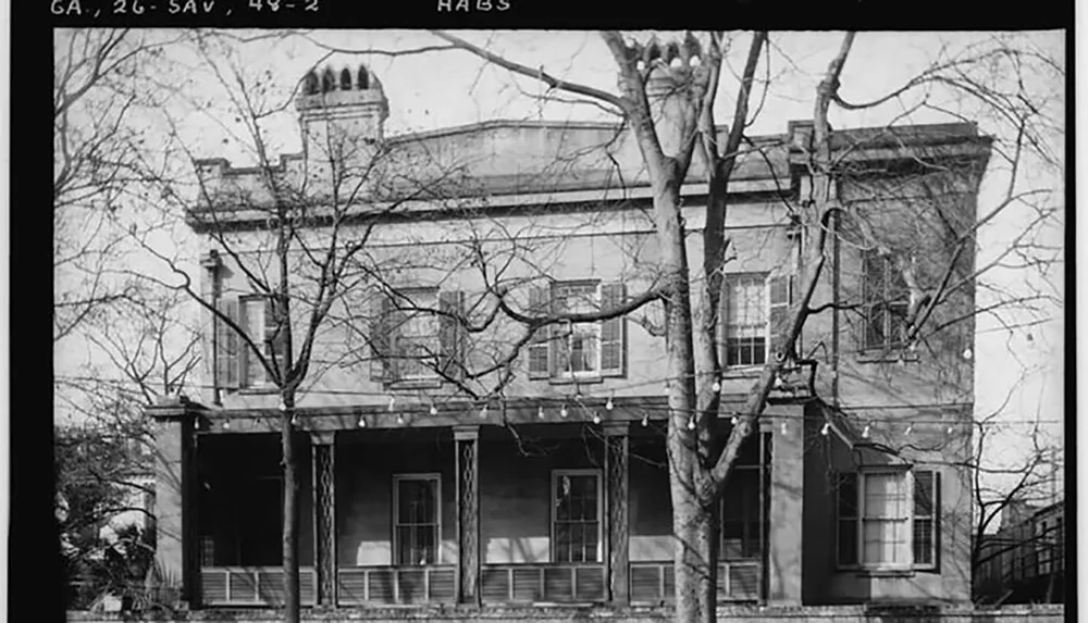 The image is a black and white photograph of a two-story historical building with a balcony surrounded by bare trees indicative of an older urban environment