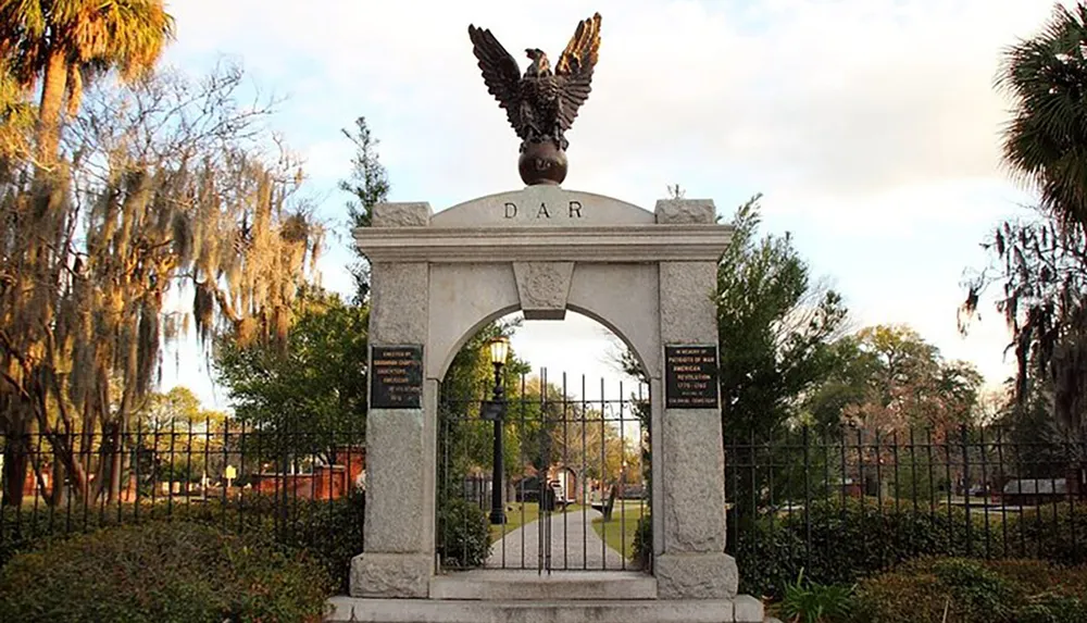 The image shows an ornate gate with the initials DAR at the top crowned by a bronze eagle sculpture flanked by trees draped with Spanish moss leading into what appears to be a park or historic site