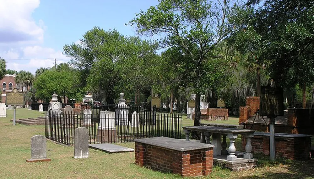 The image shows a peaceful cemetery with a variety of headstones and grave markers surrounded by trees under a blue sky