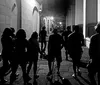 A black and white photo captures a group of people walking down a street at nighttime bathed in the ambient light from building exteriors