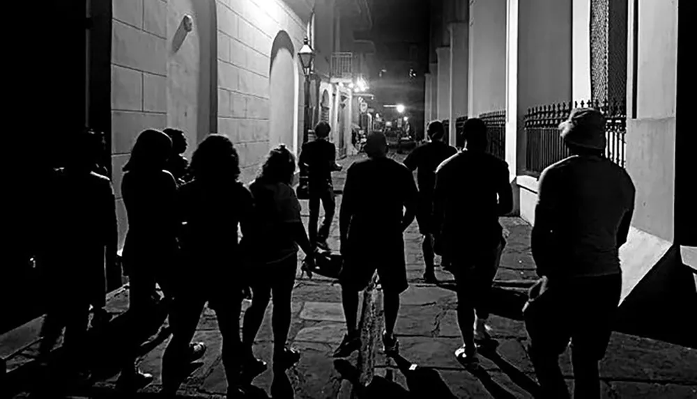 A black and white photo captures a group of people walking down a street at nighttime bathed in the ambient light from building exteriors